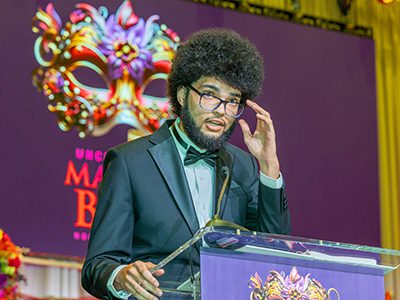 A person speaking at a podium with the Mayors' Masked Ball logo projected in the background