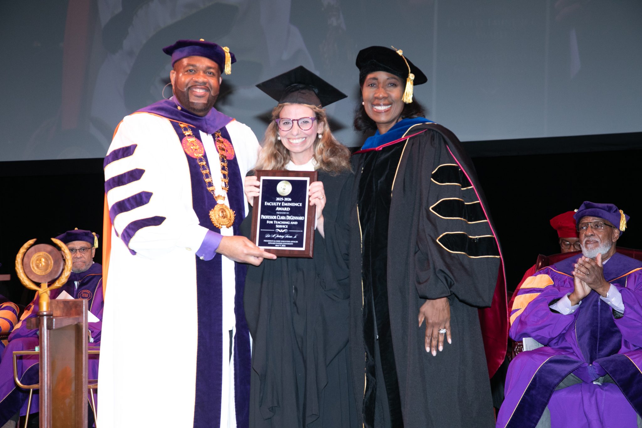 Three people in graduation attire, one holding a plaque