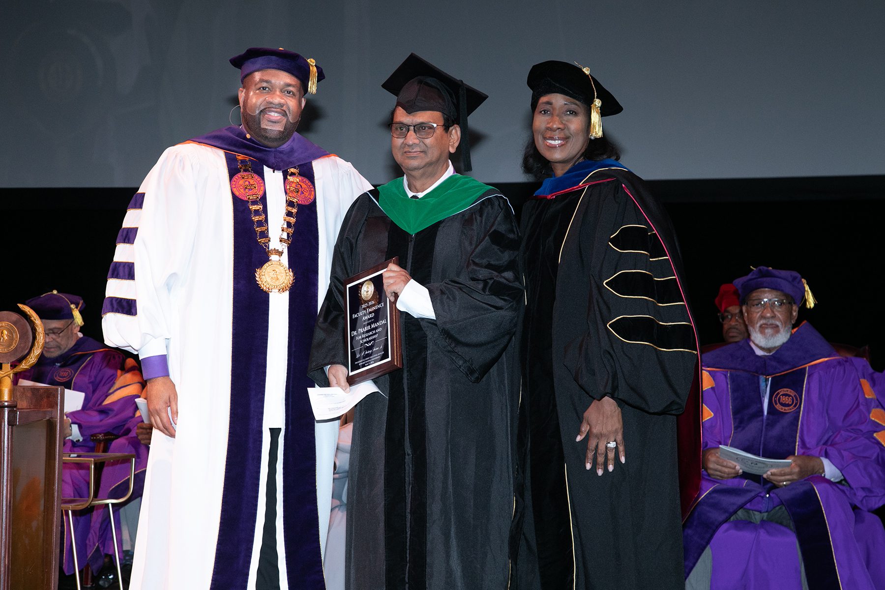 Three people in graduation attire, one holding a plaque