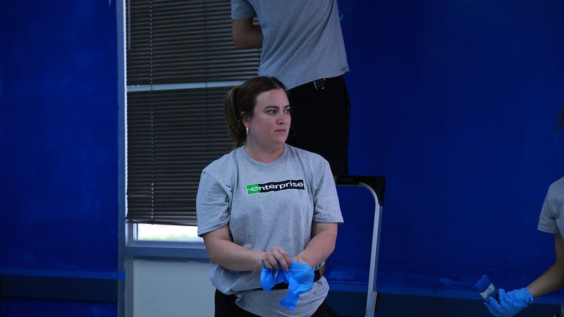 two Enterprise team members putting on gloves, another team member is standing on a ladder painting a wall