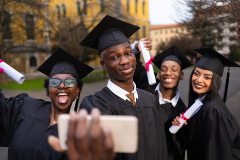 Smiling diverse graduates capturing memorable moment together, holding smartphone for group selfie after commencement