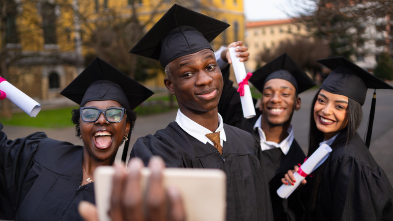 Smiling diverse graduates capturing memorable moment together, holding smartphone for group selfie after commencement