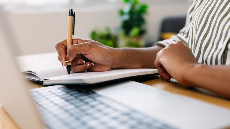 Unrecognizable businesswoman taking notes in a notebook while working on her laptop at home