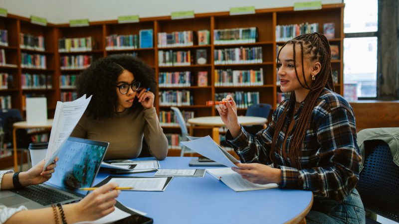 Students gathered around a table in a university library while reading, discussing and working on laptops. Authentic academic environment using both technology and real books, representing student life and group study.
