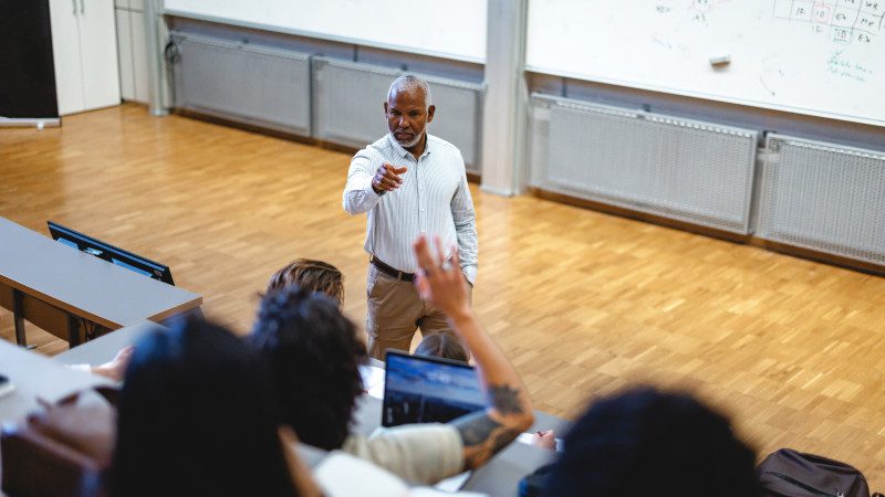Professor Pointing Toward Students While Answering A Question During University Lecture In Tiered Hall With Whiteboards And Raised Hands