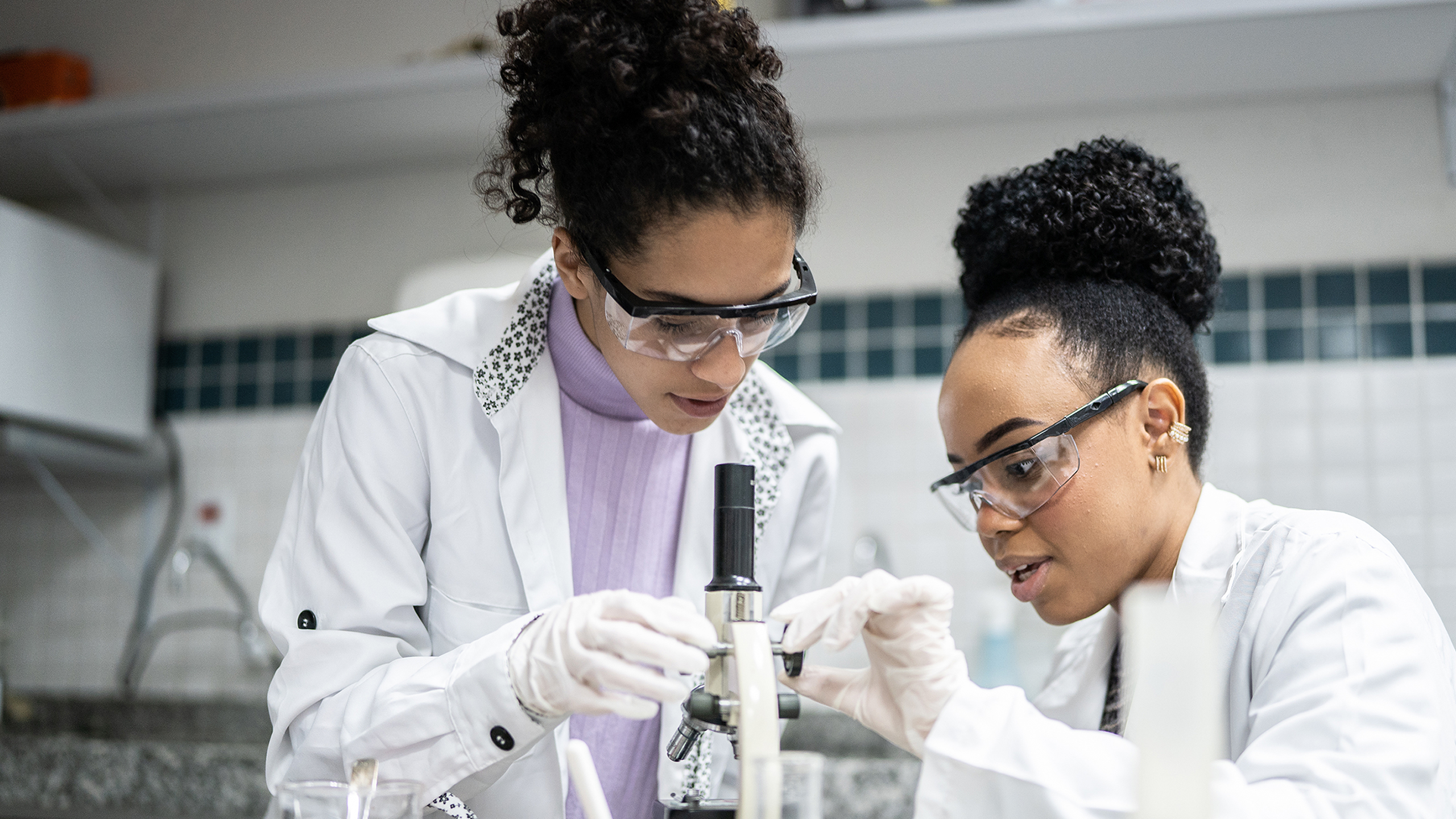 Hero Banner - two female students work together in a laboratory wearing lab coats, protective glasses and gloves