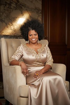 Beloved Dillard is smiling, wearing a satin taupe dress with gold embellishment, dangling earrings, and sitting in a white chair.