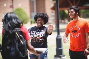 Black female student wearing a black T-shirt that says "I love my HBCU" reaches out to another female student for a hug, next to a white male student with an orange T-shirt that says "Claflin Panthers".
