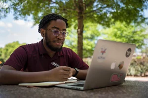 Morehouse College student holds a pen over a notebook while looking at his laptop. He sits outside under a tree.