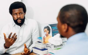A man sitting at a desk talking to another man. Photo by Ninthgrid on Unsplash.