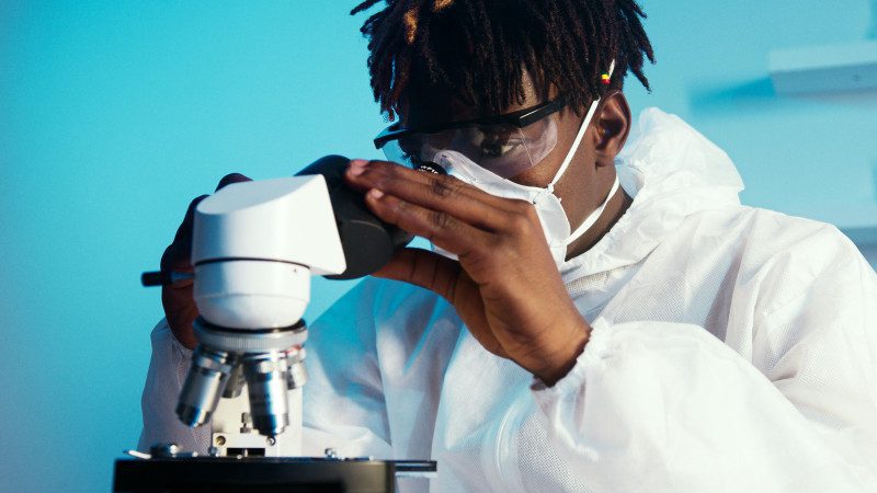 A Black researcher wearing safety glasses and a surgical mask looks through a microscope.