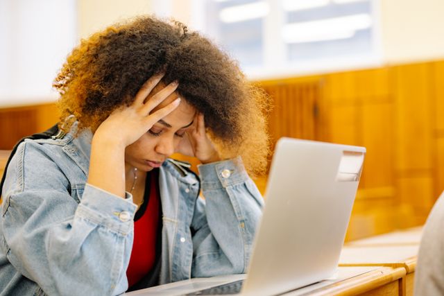A young woman experiencing a headache while studying with a laptop indoors, looking stressed and overwhelmed. Photo from Pexels/Yan Krukau.