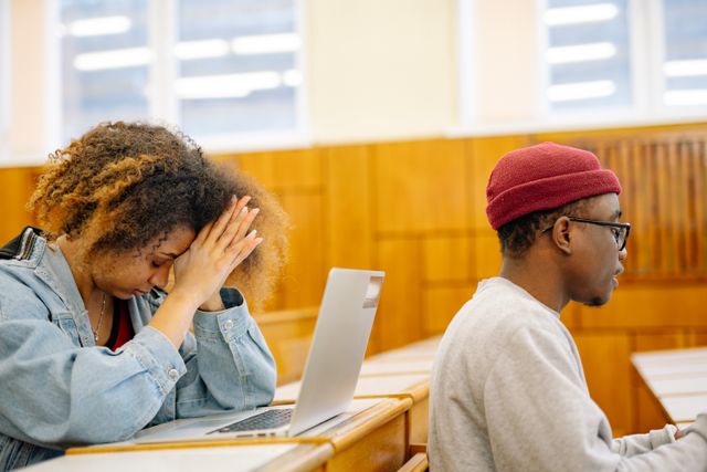 Black female college student holds her hands over her head above her laptop, looking stressed, at a desk in class, in front of her sits a Black male student facing forward.