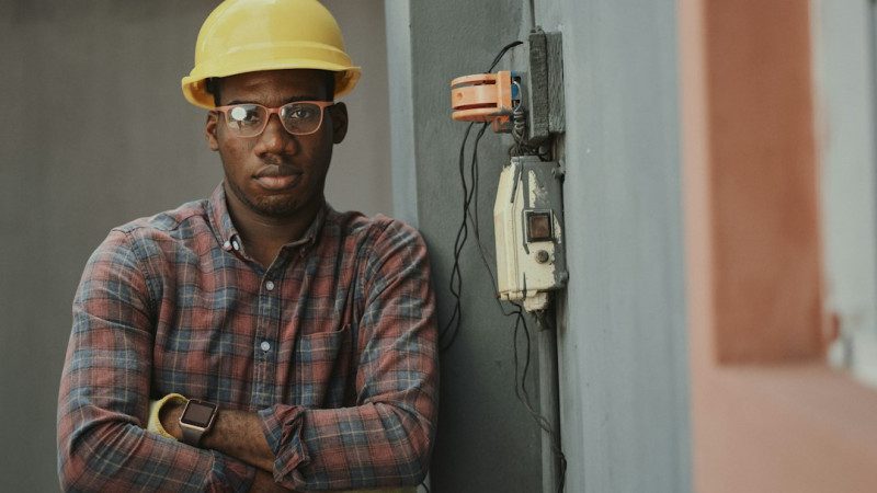 A Black man in a yellow hard hat and plaid shirt leaning against a wall. Photo by Emmanuel Ikwuegbu on Unsplash.