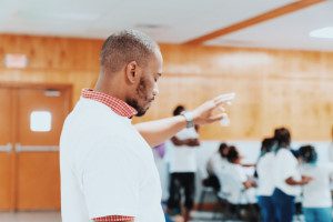 a man standing in a room with his hand raised in prayer with people in the background. Photo by Gerard Richard on Unsplash.