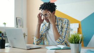 Man with dreadlocks holding head at desk with laptop. Photo by Vitaly Gariev on Unsplash.